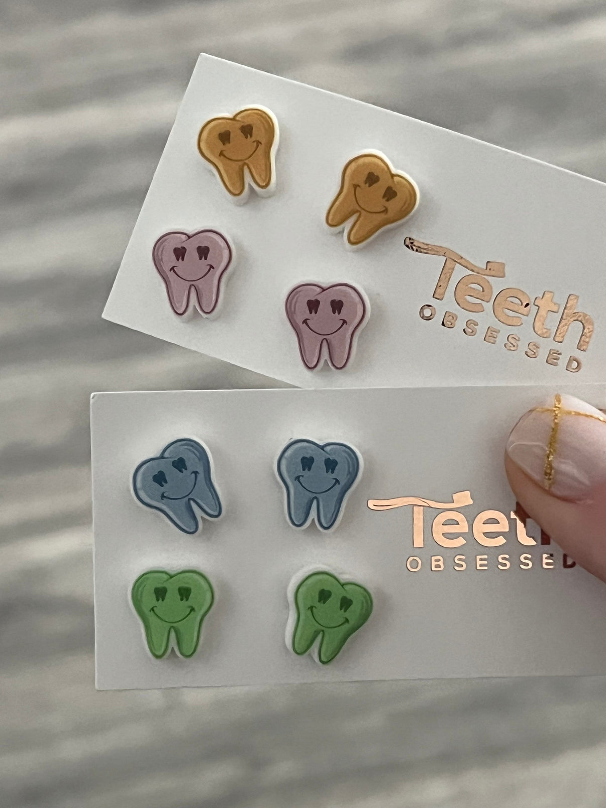 Colorful dental earrings featuring smiling teeth in green, pink, orange, and blue, designed by Teeth Obsessed.