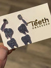 Purple Trio Dental Earrings by Teeth Obsessed featuring three small dental-themed charms, held in a hand mirror.